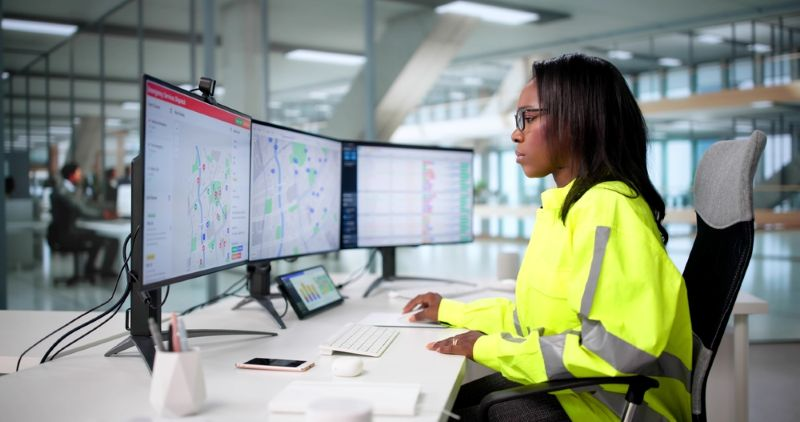 Woman in hi-vis jacket sat at a desk with three computer screens with maps on in an emergency call centre