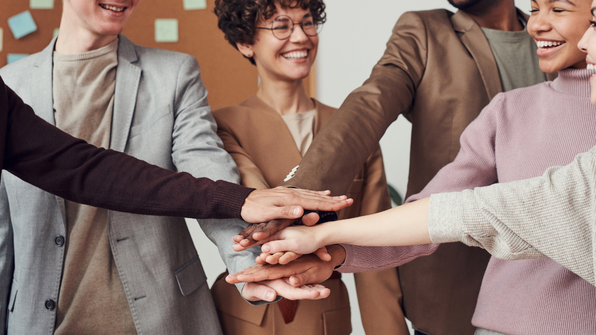 A group of people from diverse backgrounds in business suits, putting their hands on top of each others' hands and smiling