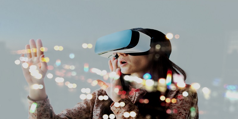 The image shows a woman wearing a virtual reality headset with her hands outstretched before her eyes. Flecks of colour and light are bursting from the headset, as she appears to gasp in awe and the world she has been transported into.