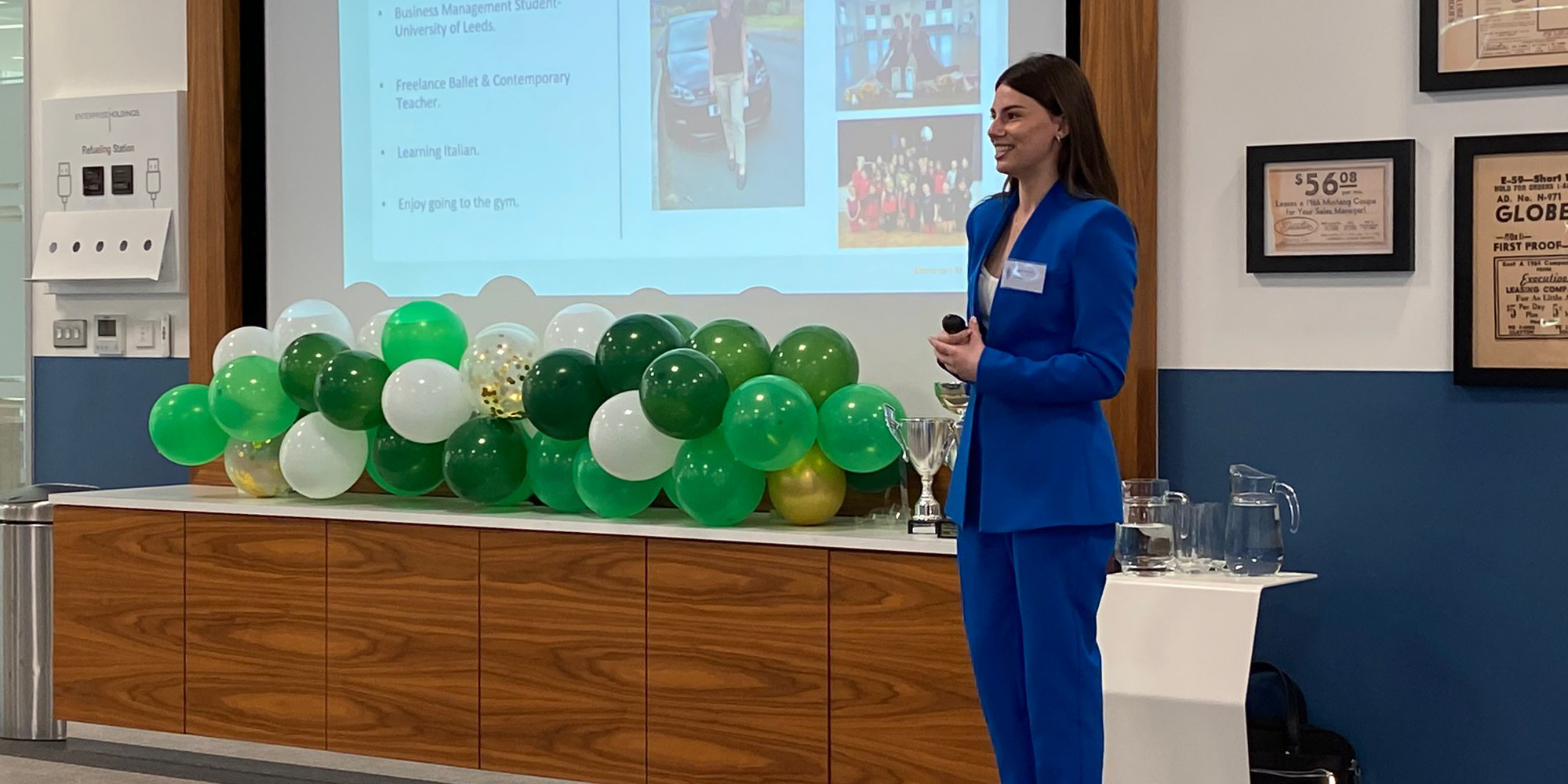 Sophie talks to crowd, whilst smiling, in front of balloons and a presentation about her business idea.