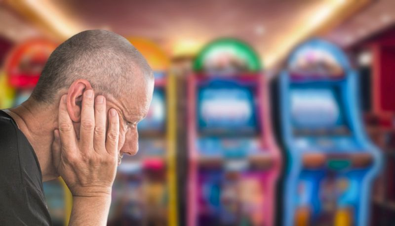 Close-up of man holding his head in his hands, with a line of gambling machines blurred out in the background