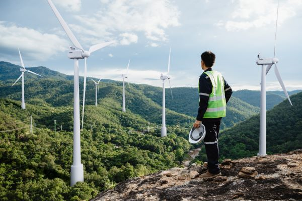 Worker wearing a hi-vis vest, holding a hard hat, standing on a mountain looking at wind turbines