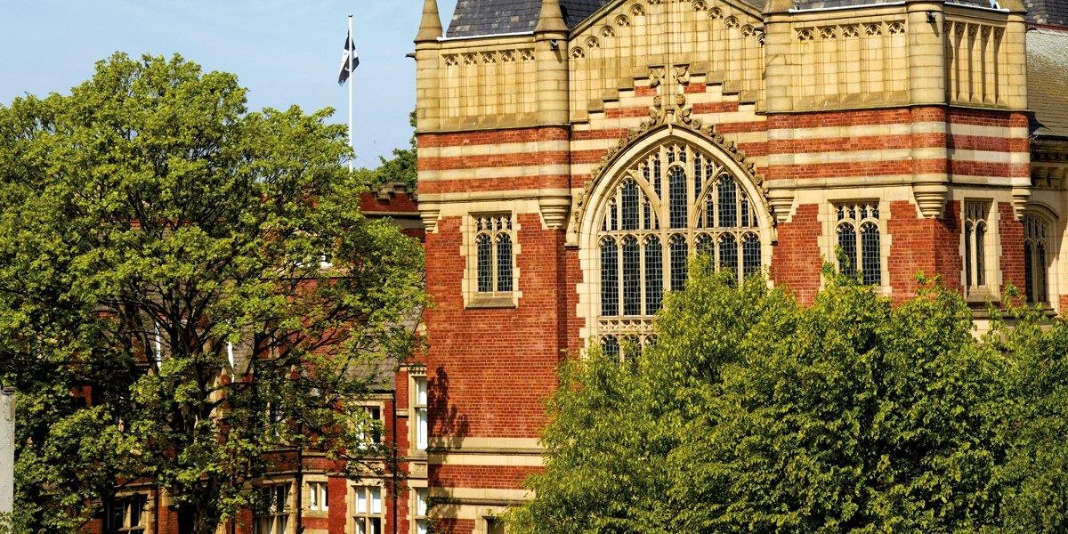 Historic University of Leeds campus building with ornate arched windows, red brick and beige stone accents, surrounded by green trees under a clear blue sky.