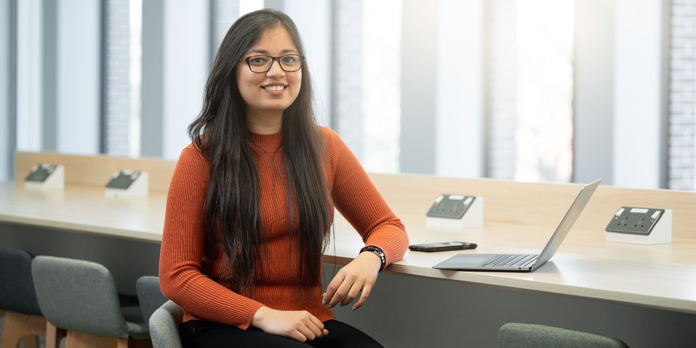 A student sat a counter work table, They are sat facing the camera with a soft smile and wearing a rust coloured long sleeve top and black trousers. They have an open laptop and a phone on the table.