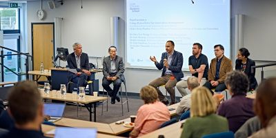 Krsto Pandza, Chee Yew Wong, Aris Theotokis, Josh Morton , Nabi Omidvar and Shahla Ghobadi presenting in front of an audience. Aris is speaking into a microphone.