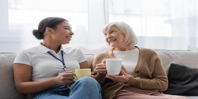 An elderly caucasian woman is sitting beside a young mixed-race care worker on a sofa in this image. They are both holding a cup of tea each and smiling, mid-conversation, at one another.