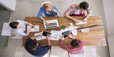 The image shows a bird's eye view of a team of five colleagues working together; laptops, tablets and papers are shown strewn across their shared working desk.