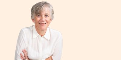 The Government Chief Scientific Adviser, Professor Dame Angela McLean, is shown off-centre to the left of the image, arms folded on a table, smiling at the camera. The background is a beige colour in keeping with the University's branding.