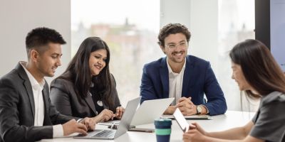 Four MBA students sitting around a desk in a collaborative suite with laptops, notes and reusable coffee cups.