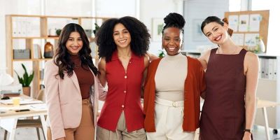 The picture shows four women of different ethnic backgrounds smiling at the camera, wearing autumnal tones of red and brown, while an open-plan office space is visible in the background.