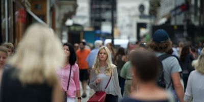 The image shows a young, blonde, white woman walking through an urban metropolis with people of different ages, races and nationalities.
