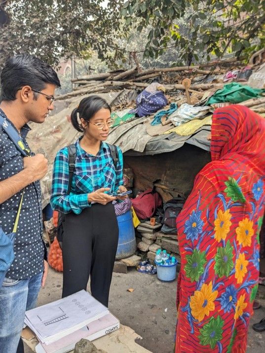 Two survey interviewers stand beside a makeshift home, using a phone and papers while speaking with a resident