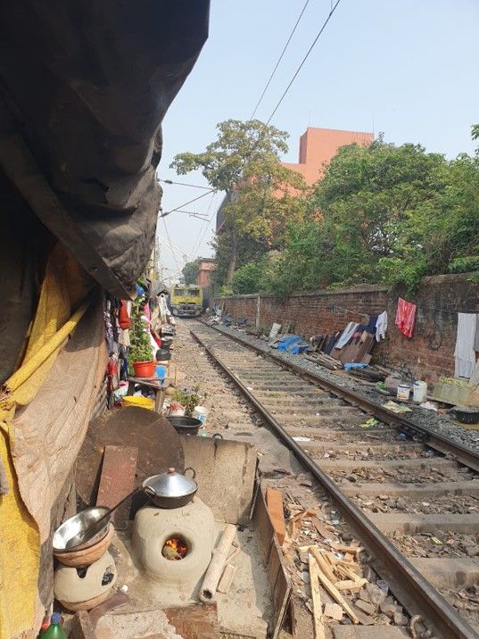 Kolkata slum - Railway track passing beside tightly packed informal homes, with a small stove and utensils in the foreground and a train approaching in the distance.