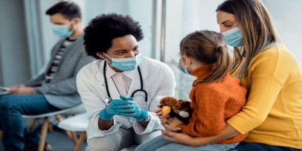 The image shows a Black doctor interacting with a young girl and her mother. The child is sitting on the mother's lap; the doctor wears a mask and gloves, white scrubs and leans forward with smiling eyes to engage with her patient.