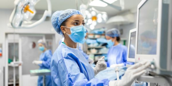 Healthcare professional in surgical scrubs, mask and gloves using a medical monitor in an operating theatre, with other staff and equipment in the background