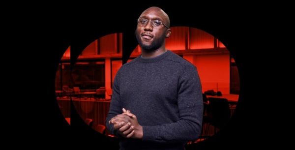 Man clasping hands at his waist whilst looking confidently into camera against a black and red backdrop