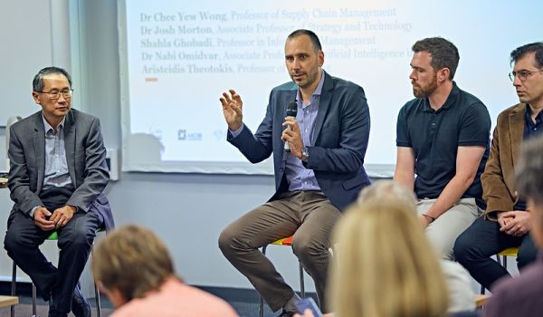 Chee Wong, Aris Theotokis, Josh Morton and Nabi Omidvar sat in front of an audience with a presentation screen behind them. Aris is speaking into a microphone.