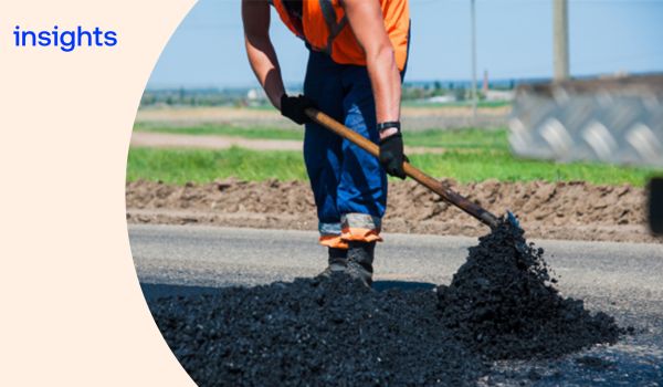 A worker in high‑visibility clothing uses a tool to spread fresh asphalt across a road