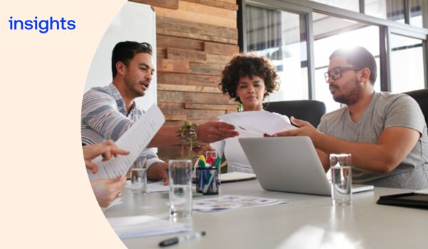 Three office workers sat around a table, going through some papers with a laptop in front of them