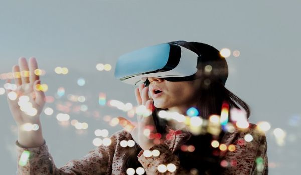 The image shows a woman wearing a virtual reality headset with her hands outstretched before her eyes. Flecks of colour and light are bursting from the headset, as she appears to gasp in awe and the world she has been transported into.