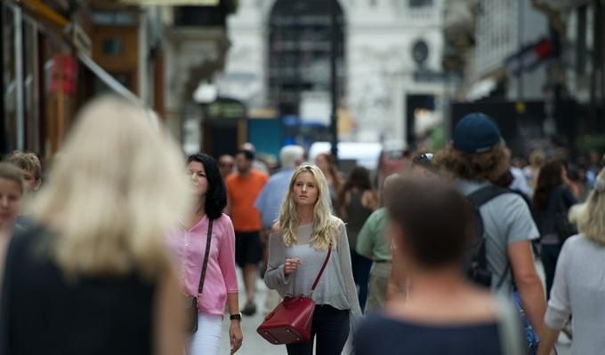The image shows a young, blonde, white woman walking through an urban metropolis with people of different ages, races and nationalities.