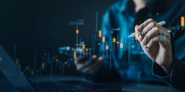 Person using a stylus and smartphone in front of a digital financial dashboard, with candlestick charts and market data visualisations overlaid on a dark blue background, representing fintech technology.