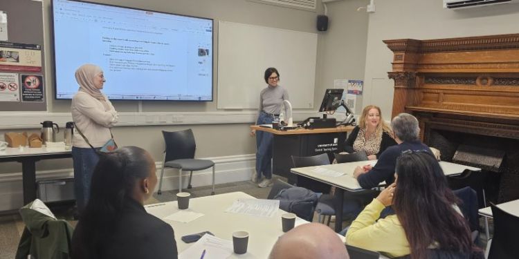 Two people stood at the front of a room in front of a presentation, smiling at someone who is sat down, talking to other people, in a classroom setting