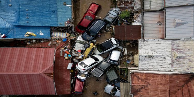 The image shows a bird's eye view of a flooded area with a pile of multi-coloured vehicles in the middle, surrounded by the roofs of an urban area.
