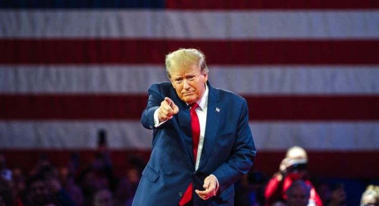 Donald Trump, pointing forwards, stood in front of an American flag