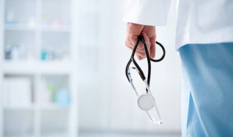 A hand holding a stethoscope in what looks to be a medical room.