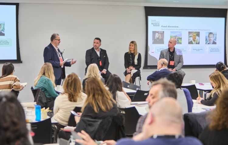 David Loseby speaking into a microphone next to the panel members who are sat in front of an audience. From left to right, the panel is: Stefan Jastak, Caroline Hinchcliffe and Matthew Rumble