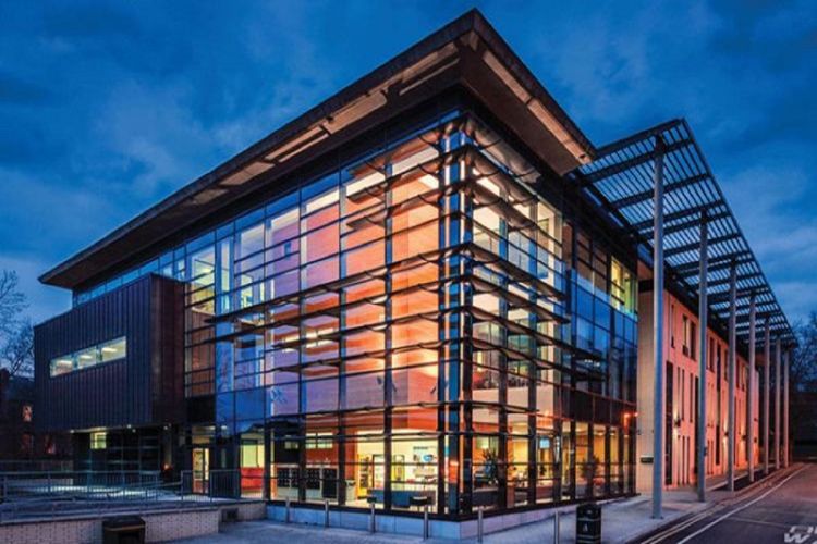 The image shows the School of Law's Liberty Building with bright lights on inside at the foreground, which contrasts with the early evening sky behind the building.