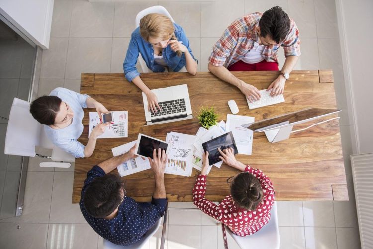 The image shows a bird's eye view of a team of five colleagues working together; laptops, tablets and papers are shown strewn across their shared working desk.