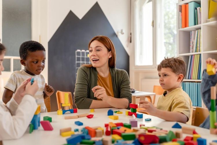 The image shows a red-headed early years worker sitting between two children, surrounded by multi-coloured building blocks, engaged in play. The worker and children show a degree of ethnic and representative diversity. 