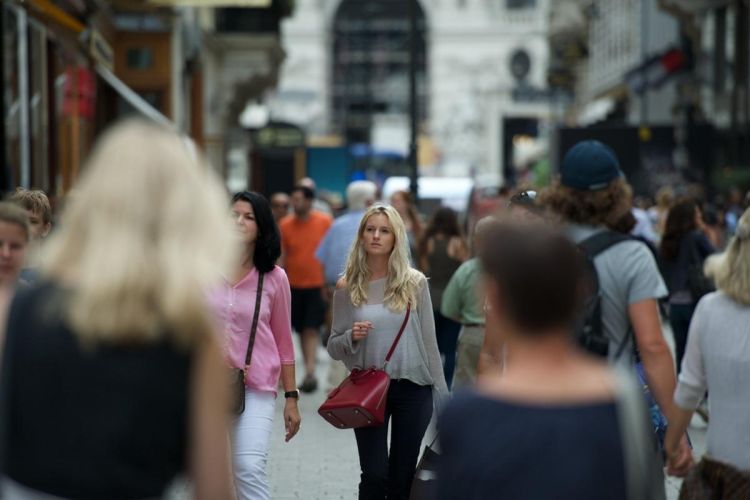 The image shows a young, blonde, white woman walking through an urban metropolis with others of mixed ages, races and nationalities.