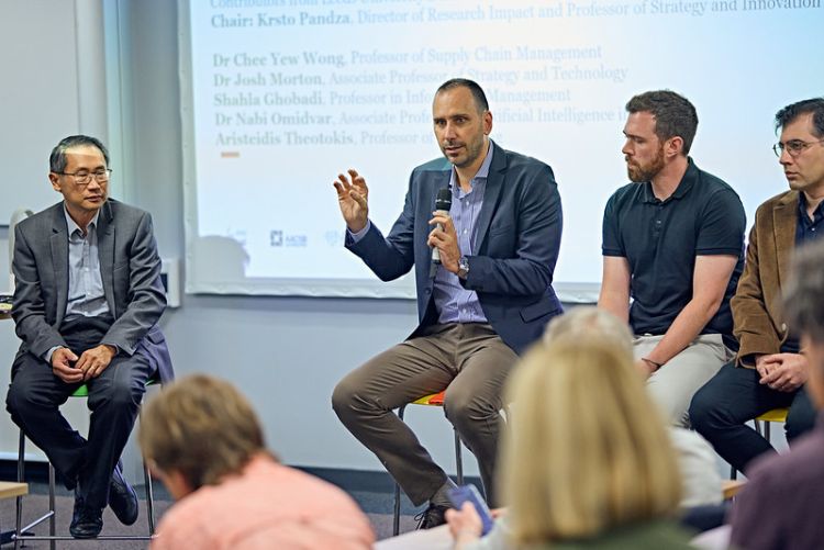 Chee Wong, Aris Theotokis, Josh Morton and Nabi Omidvar sat in front of an audience with a presentation screen behind them. Aris is speaking into a microphone.