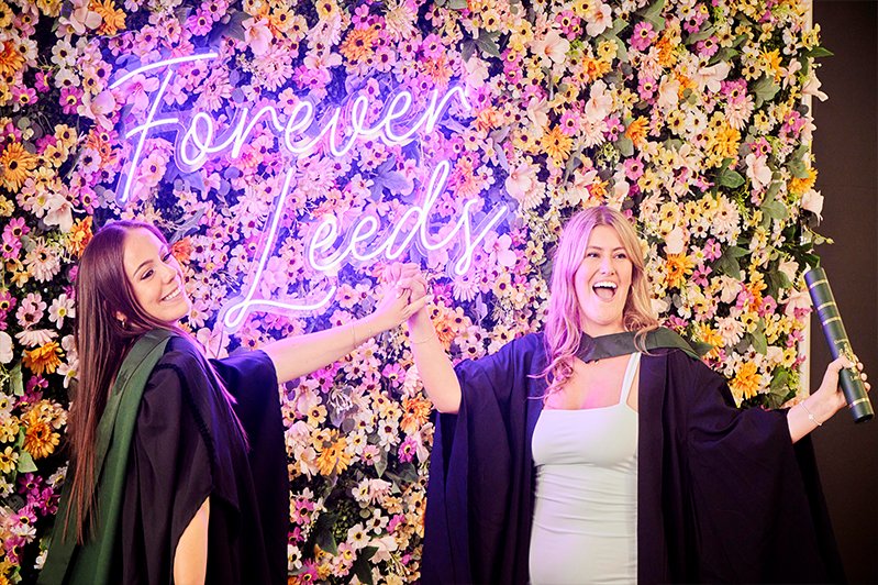 Two graduating female students, smiling and holding hands raised in celebration, stand in front of a vibrant flower wall with a neon pink sign that reads "Forever Leeds."
