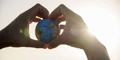 Closeup of hands with a world globe in their hands forming a heart against the sky and a ray of sun showing up on the right